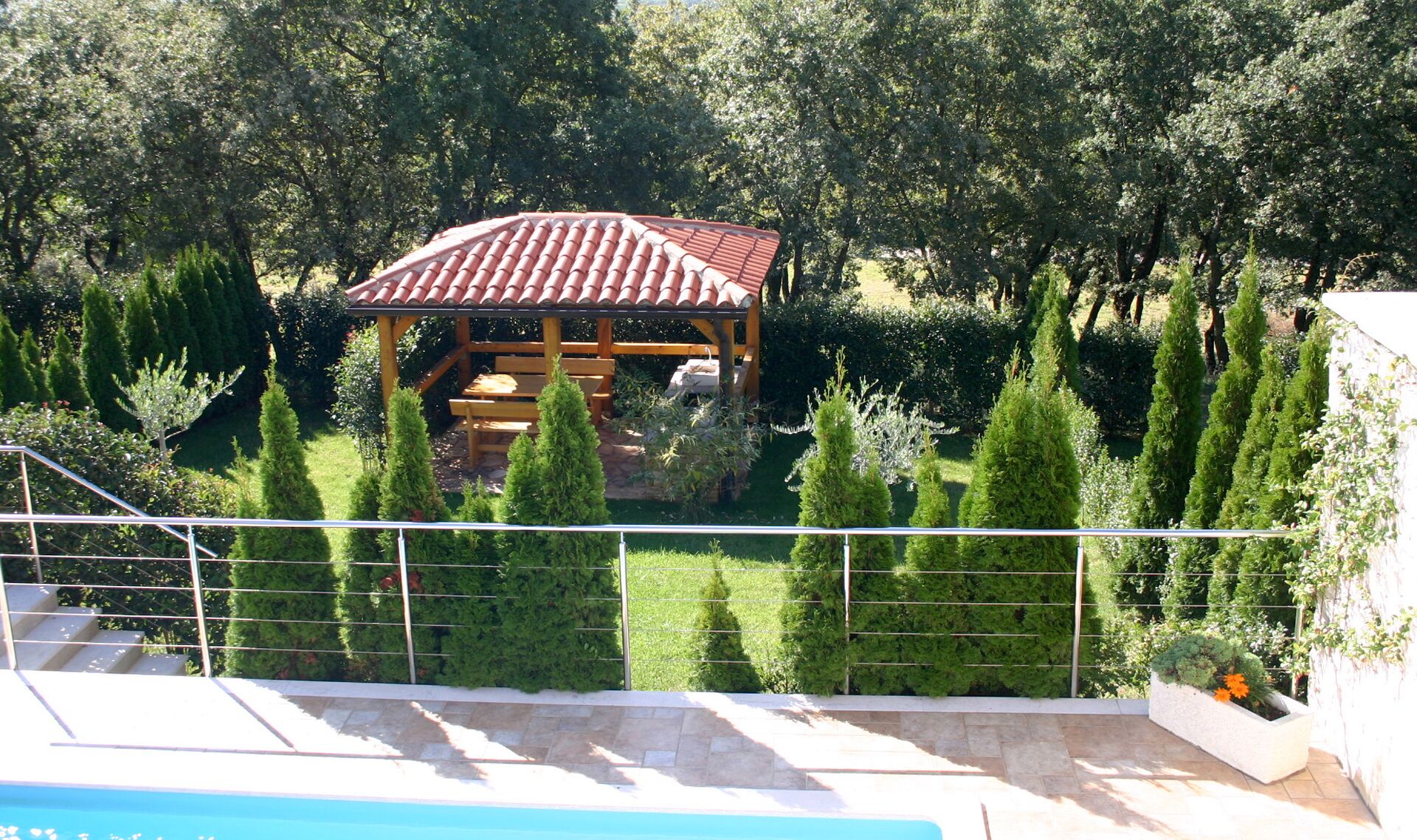 A gazebo with a tiled roof, surrounded by neatly trimmed hedges and trees, overlooking a swimming pool area.