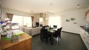 Ett modernt vardagsrum med en soffa, ett matbord med stolar, en köksdel och ett stort fönster med gardiner.