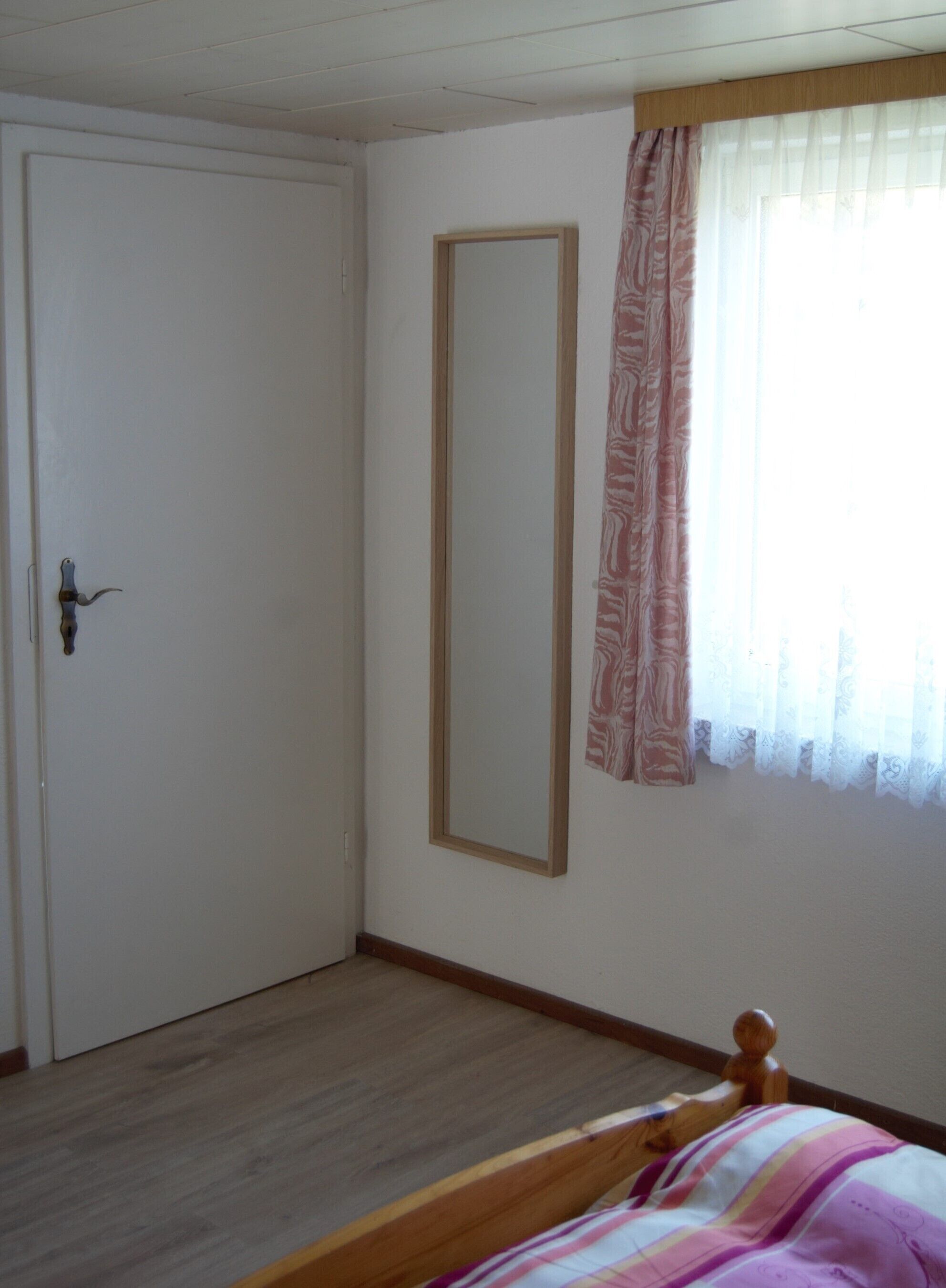A bedroom with a wooden bed, a wardrobe, a mirror, and a window with curtains.