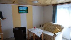 Una sala de estar con un sofá, una mesa con mantel, una silla, un cuadro en la pared y una ventana con cortinas.