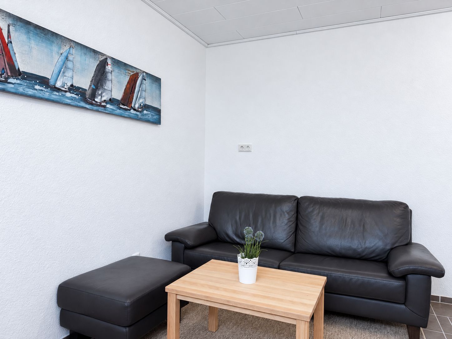 Un salon avec un canapé en cuir noir, une table basse en bois et un tableau représentant des voiliers accroché au mur.
