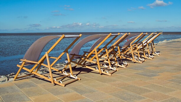 Eine Reihe aus Holzliegen mit weißen Kissen, aufgemacht entlang einer gefliesten Promenade am Meer.