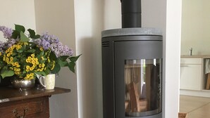 A modern wood-burning stove with a glass door, positioned against a white wall in a room with a wooden dresser and a vase of flowers. - Villa holiday Stintenburg island in the Biosphere (Zarrentin am Schaalsee OT Stintenburg)