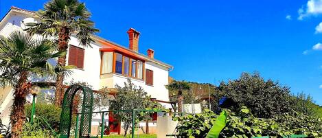 A white building with red shutters and a red roof, surrounded by lush greenery and a clear blue sky.