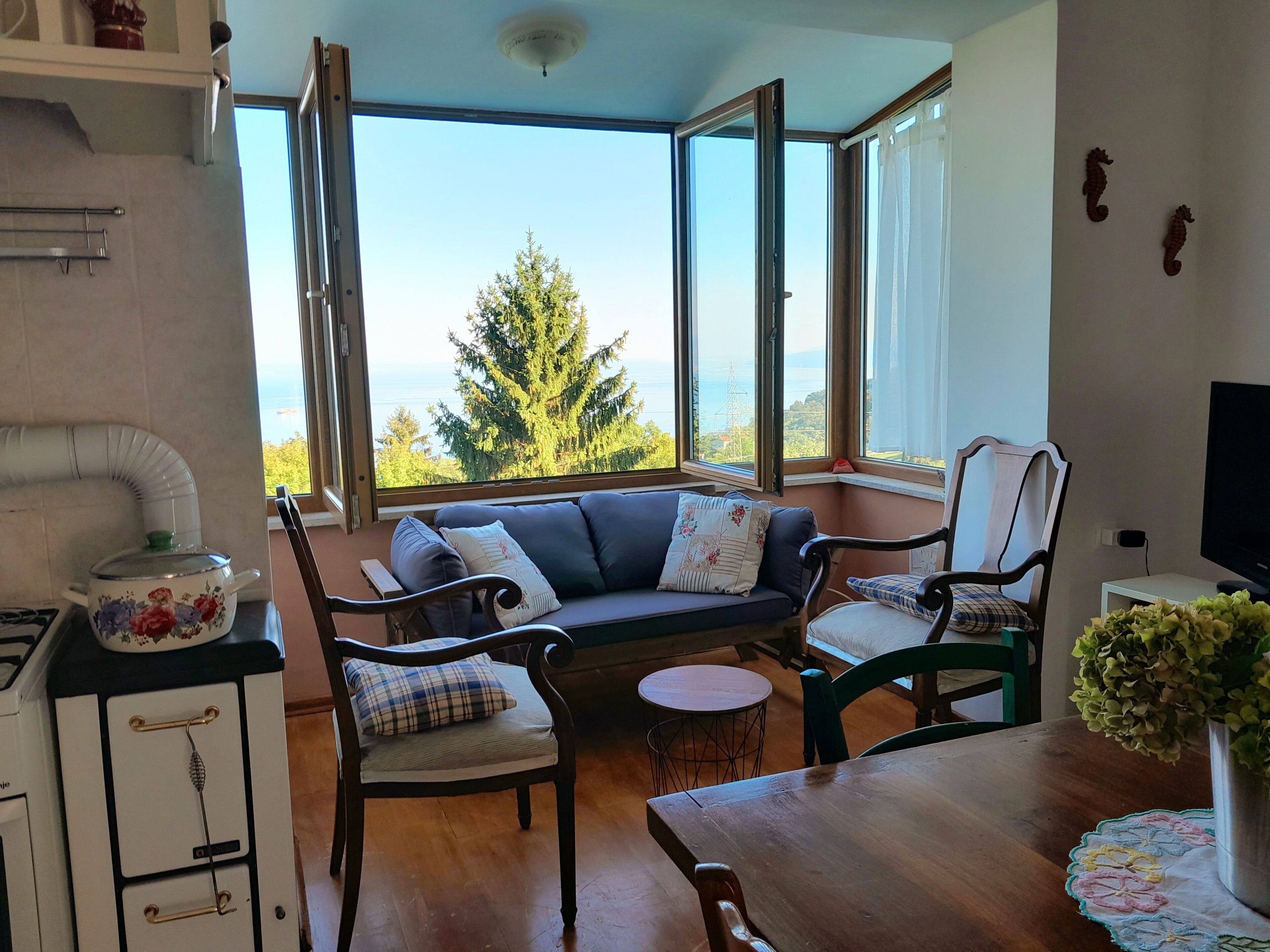 Une pièce confortable avec un canapé, des fauteuils, une petite table et un téléviseur. De grandes fenêtres offrent une vue sur les arbres et la mer.