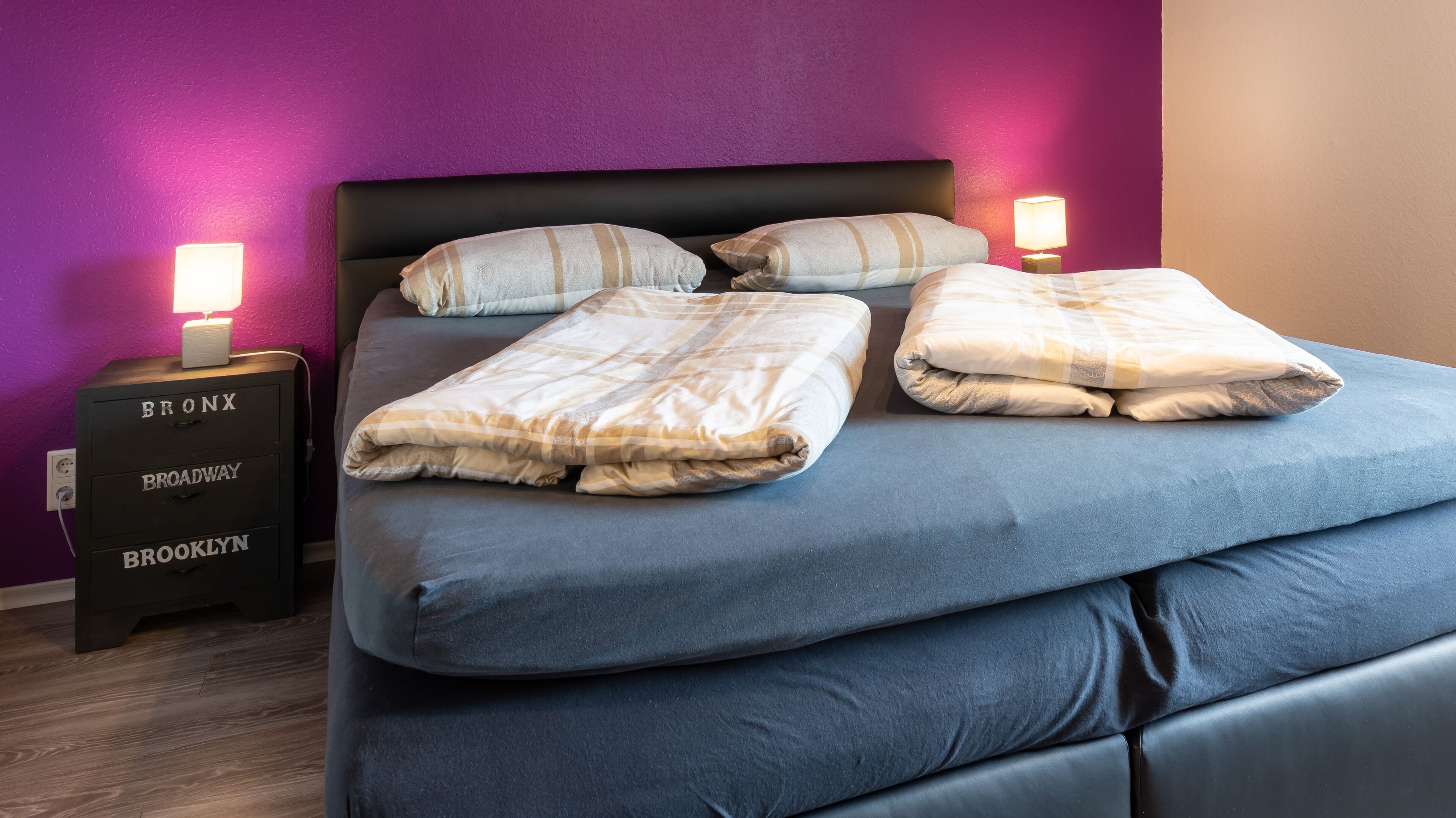 A bed with a blue mattress and two pillows, a bedside table with a lamp, and a dresser with drawers labeled 'Bronx,' 'Broadway,' and 'Brooklyn.'