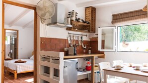 A compact kitchen with a refrigerator, a dining table with chairs, and a bed in the adjacent room. - Tasteful house, 2 double rooms, 2 bathrooms, large terrace, garden with outdoor shower,  (Es Pujols)