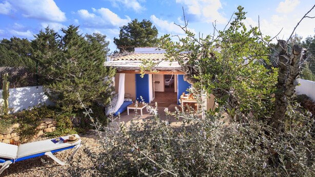 A small house with a tiled roof, a patio with a table and chairs, and a garden with various plants and a tree. - Tasteful house, 2 double rooms, 2 bathrooms, large terrace, garden with outdoor shower,  (Es Pujols)