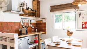 A compact kitchen with a refrigerator, wooden shelves, hanging utensils, and a dining area with chairs and a table. - Tasteful house, 2 double rooms, 2 bathrooms, large terrace, garden with outdoor shower,  (Es Pujols)