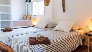 A neatly made bed with striped bedding and a brown towel, a bedside table with a lamp, and a window with a view of the outside. - Tasteful house, 2 double rooms, 2 bathrooms, large terrace, garden with outdoor shower,  (Es Pujols)