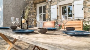 Une table de jardin rustique, dressée pour un repas en plein air, avec des verres à vin, des bols et des assiettes, placée devant un bâtiment en pierre agrémenté de meubles en bois.