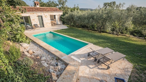 Une piscine rectangulaire entourée d’une terrasse en pierre, un petit bâtiment avec un toit en tuiles et une zone herbeuse agrémentée d’arbres et d’arbustes.