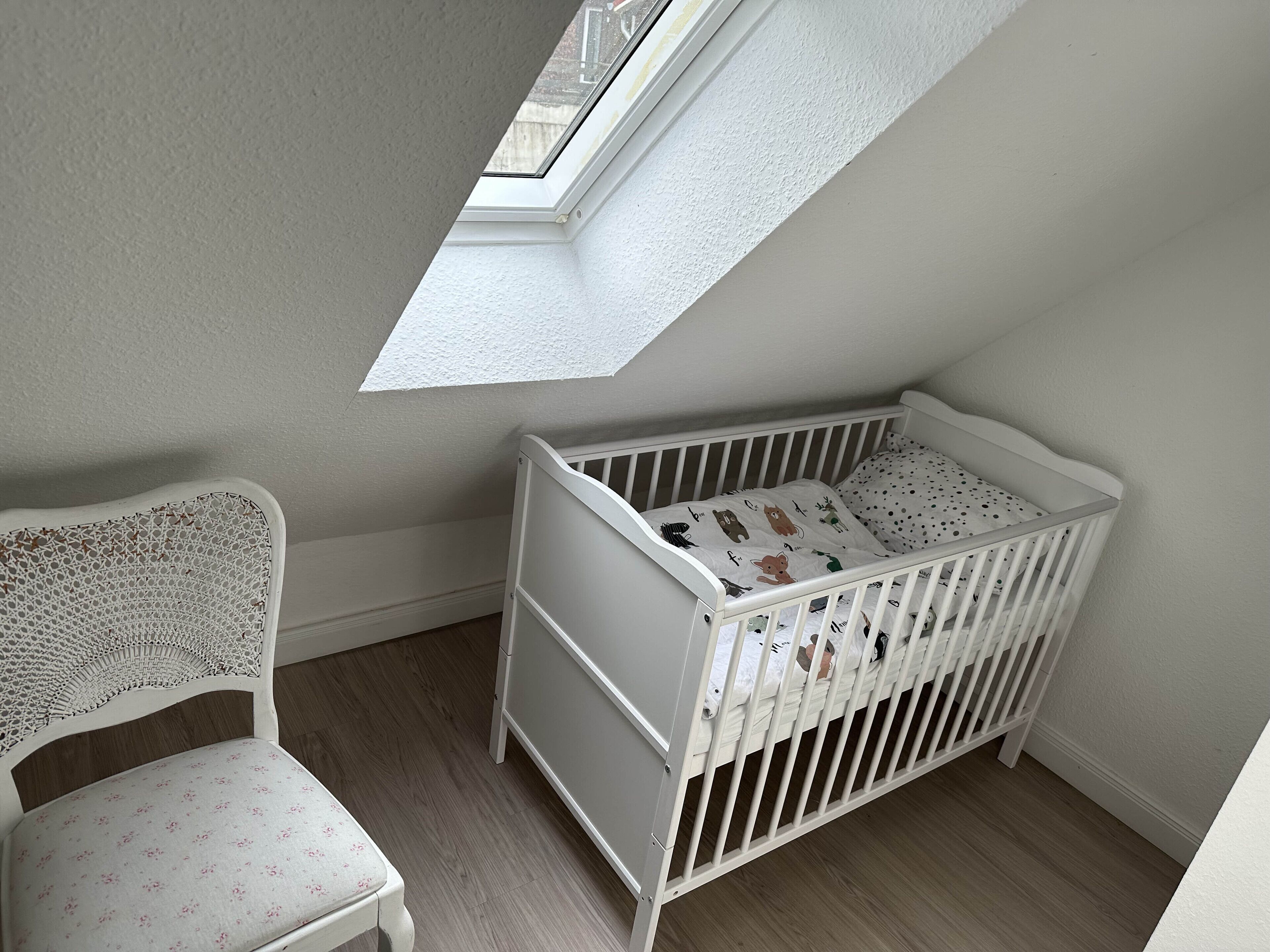 Una cuna blanca con colchón de lunares, una silla blanca con estampado floral y un tragaluz en una habitación con paredes de colores claros y suelo de madera.