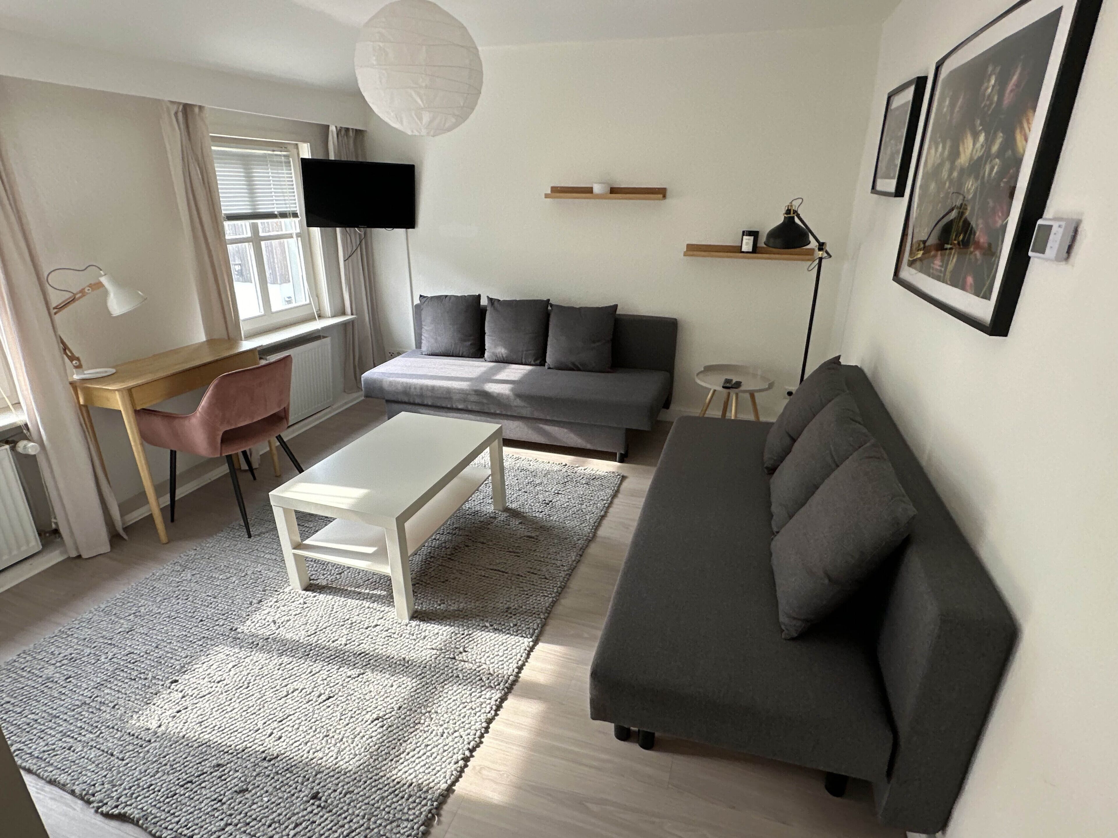 Een moderne woonkamer met een grijze bank, een witte salontafel, een houten bureau en een aan de muur bevestigde televisie.