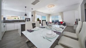 Ein moderner Essbereich mit einem weißen Tisch, gedeckt für vier Personen, einer Küche im Hintergrund und Blick auf das Meer durch große Fenster.