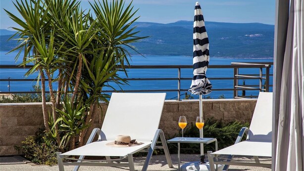 Eine Terrasse mit zwei weißen Liegestühlen, einem gestreiften Sonnenschirm und einem Tisch mit zwei Gläsern Orangensaft.