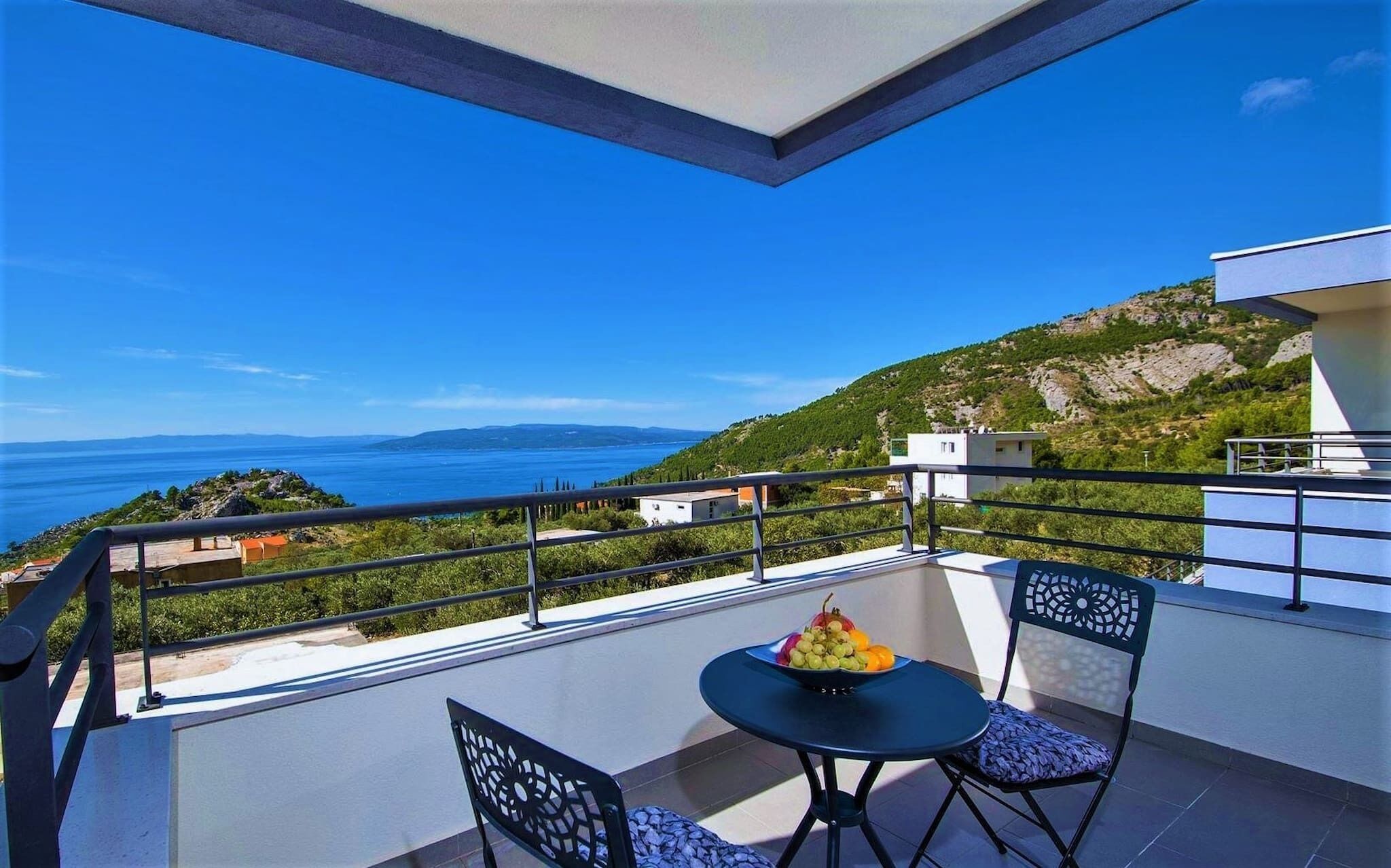 A balcony with a table and chairs, a bowl of fruit, and a view of the sea and hills.