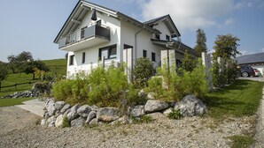Una casa moderna a due piani con un balcone, immersa in un giardino con diverse piante e rocce.