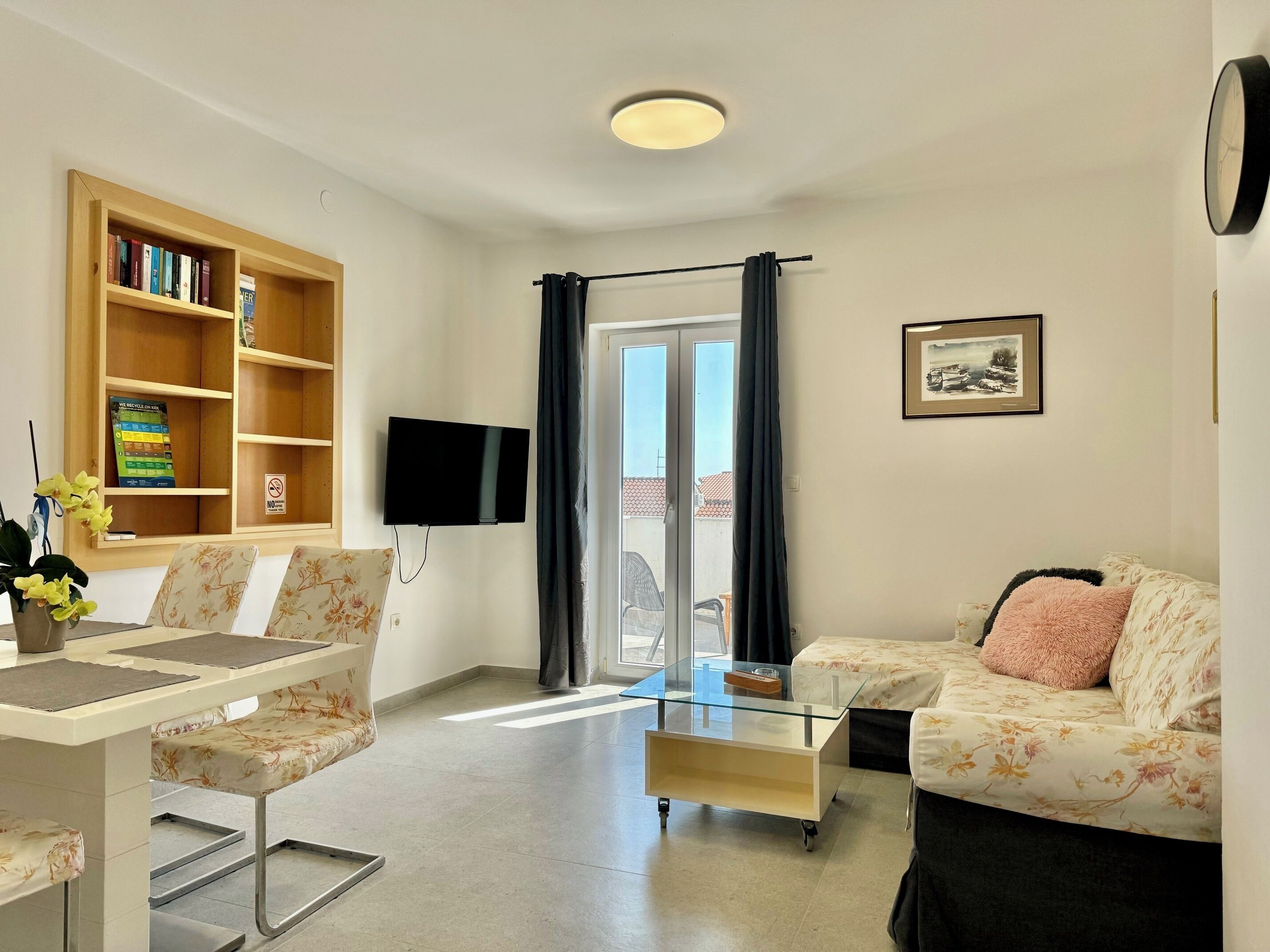 A living room with a sofa, a coffee table, a TV mounted on the wall, a bookshelf, and a glass door leading outside.
