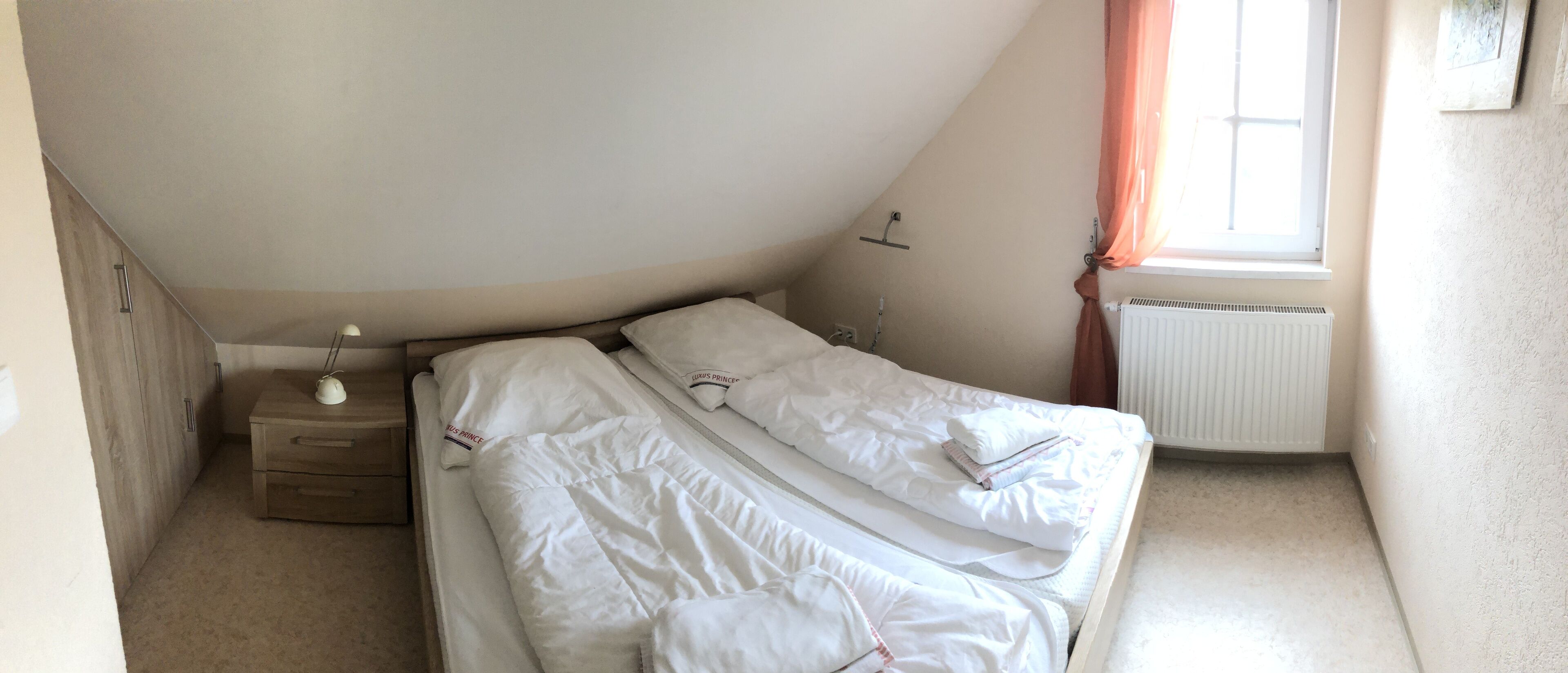 A small, single-bed room with a sloped ceiling, a bedside table with a lamp, a radiator, and a window with curtains.