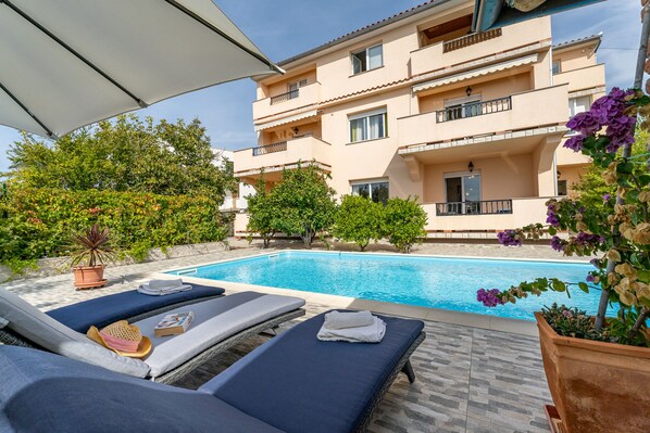 A poolside area with lounge chairs, a pool, and a building with balconies. - APARTMENTS JURIĆ WITH SWIMMING POOL, 15 MIN. TO THE CENTER, 5 MIN. TO THE BEACH (RAB)