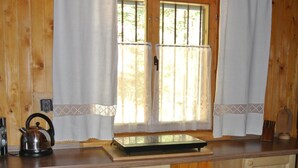 A kitchen with wooden cabinets, a small wooden stool, a kettle, and a window with curtains. - Comfortable, romantic vacation home right at a national park (Jakubovany)