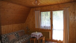 A cozy cabin interior with a wooden ceiling, a sofa with patterned cushions, a small table with a white tablecloth, and a large window with curtains. - Comfortable, romantic vacation home right at a national park (Jakubovany)