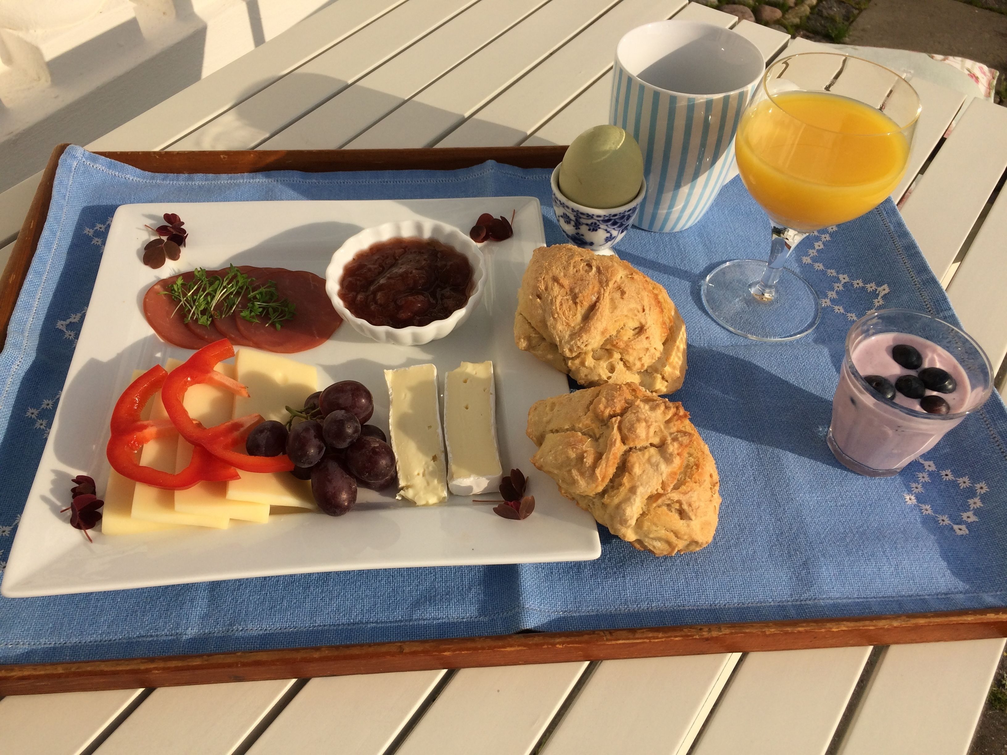 Ein Tablett mit Käse, Trauben, roten Paprika, einer Marmeladenschale, Keksen und einem Glas Orangensaft auf einem blauen Tuch.