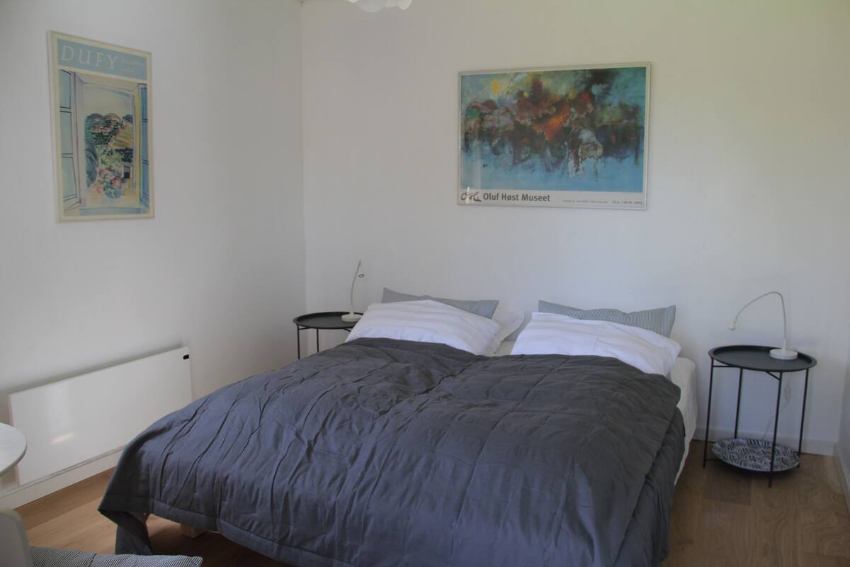 A bedroom with a bed, two bedside tables, a nightstand, a radiator, and a painting on the wall.