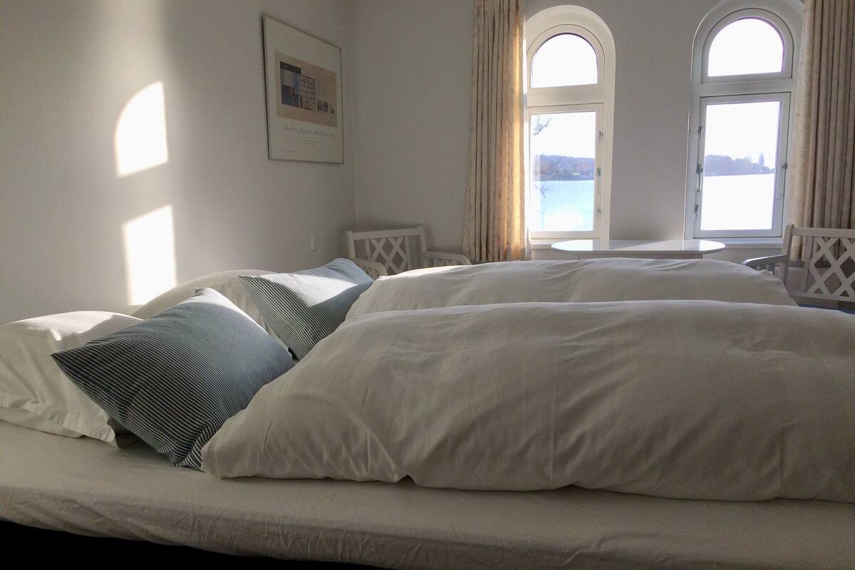 A bedroom with a large bed, two bedside tables, a window with curtains, and a framed picture on the wall.