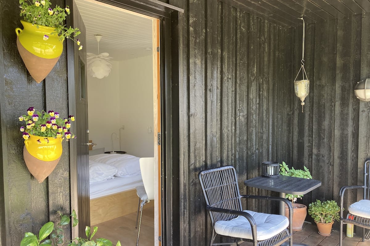 A room with a bed, a chair, a table, and hanging planters with flowers.