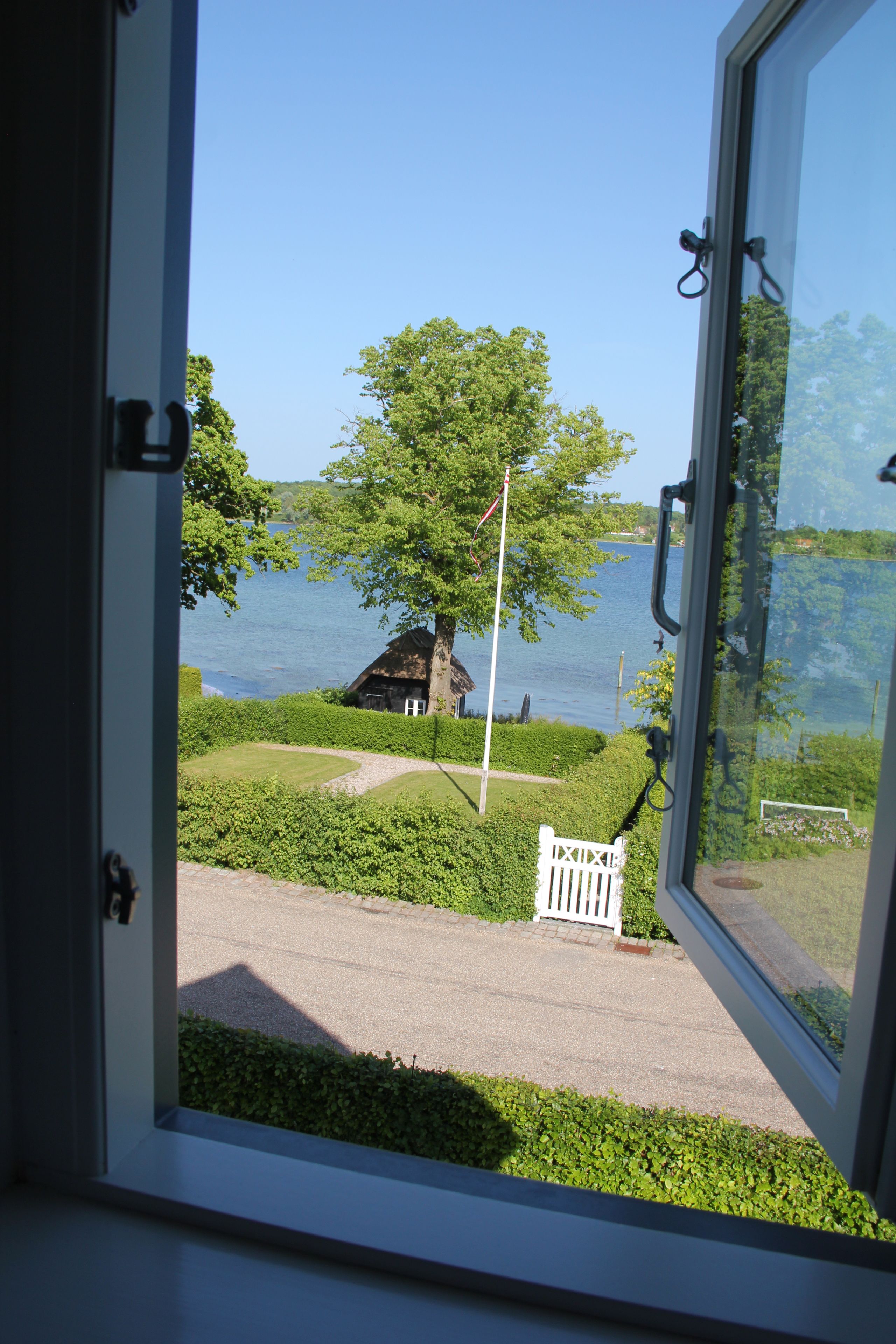 A view through a window of a house by a lake, with a flagpole and a white fence visible.