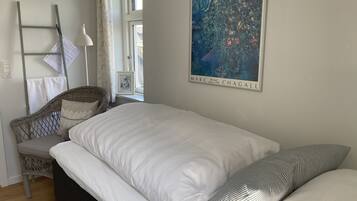 Ett sovrum med en säng, en flätad stol, en golvlampa, en inramad tavla och ett fönster med gardiner.