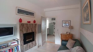 Een woonkamer met een televisie, boekenkasten, een open haard, een bank en een deur naar een keuken.