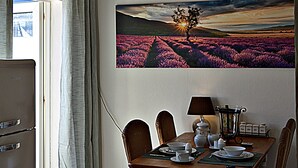 Een eethoek met een tafel voor vier personen, een schilderij van een zonsondergang over een veld, en een plafondlamp.