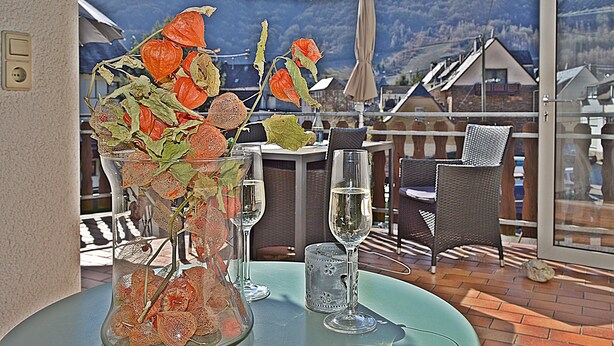 Een tafel met een vaas gedroogde bloemen, een glas wijn en een glas water op een balkon met uitzicht op een berglandschap.