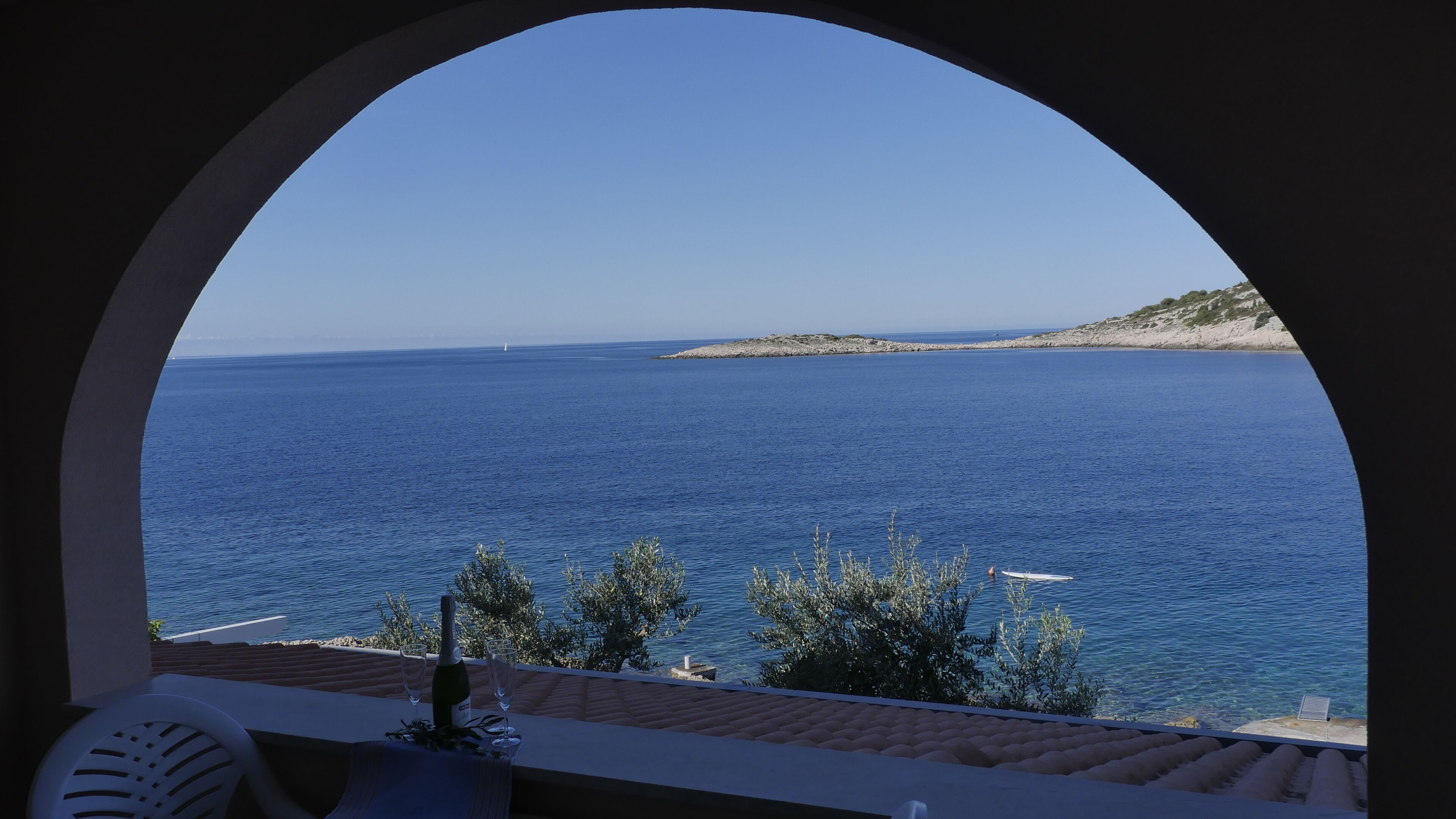 A view through an arched window of a coastal scene with a clear blue sea, a distant island, and a table with a bottle and glasses.
