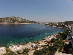 Une ville côtière aux eaux d’un bleu limpide, avec des bateaux et une plage aménagée avec des parasols et des chaises longues.