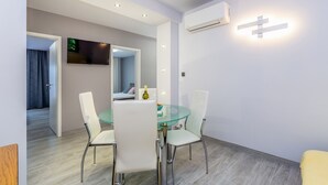Un espace repas moderne avec une table en verre et des chaises blanches, un téléviseur à écran plat fixé au mur et un éclairage encastré au plafond.
