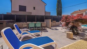 Une terrasse avec des chaises longues, une table entourée de chaises, une piscine et un bâtiment doté d’un toit en pente.