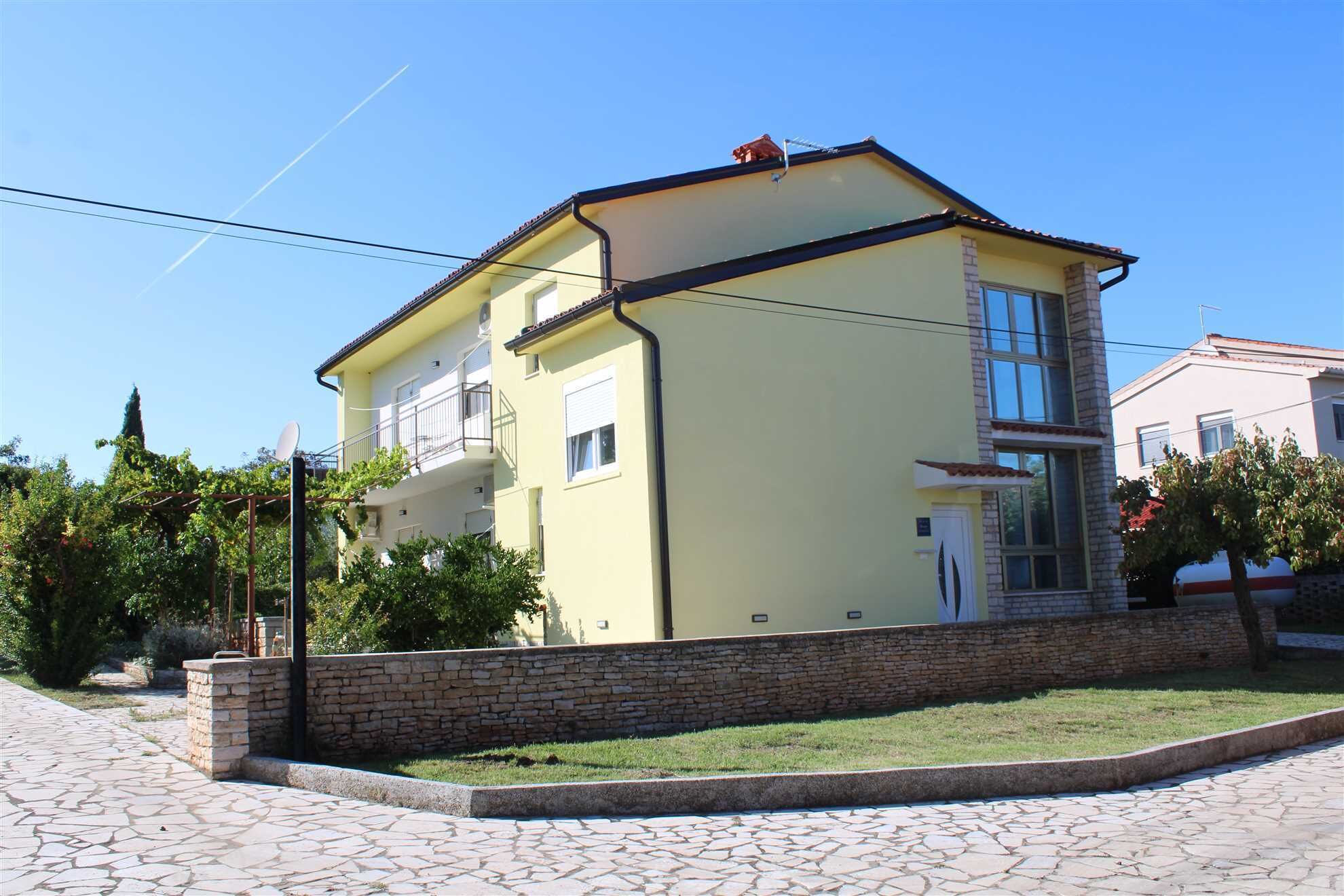 Um edifício de dois andares com fachada amarela, uma parede de pedra à frente e um espaço pavimentado com caminho de pedra.