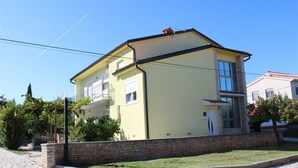 A two-story building with a yellow facade, a stone wall in front, and a paved area with a stone pathway. - Cozy 3-Bedroom Haven, Perfect for 5, With Parking Space (Ližnjan)