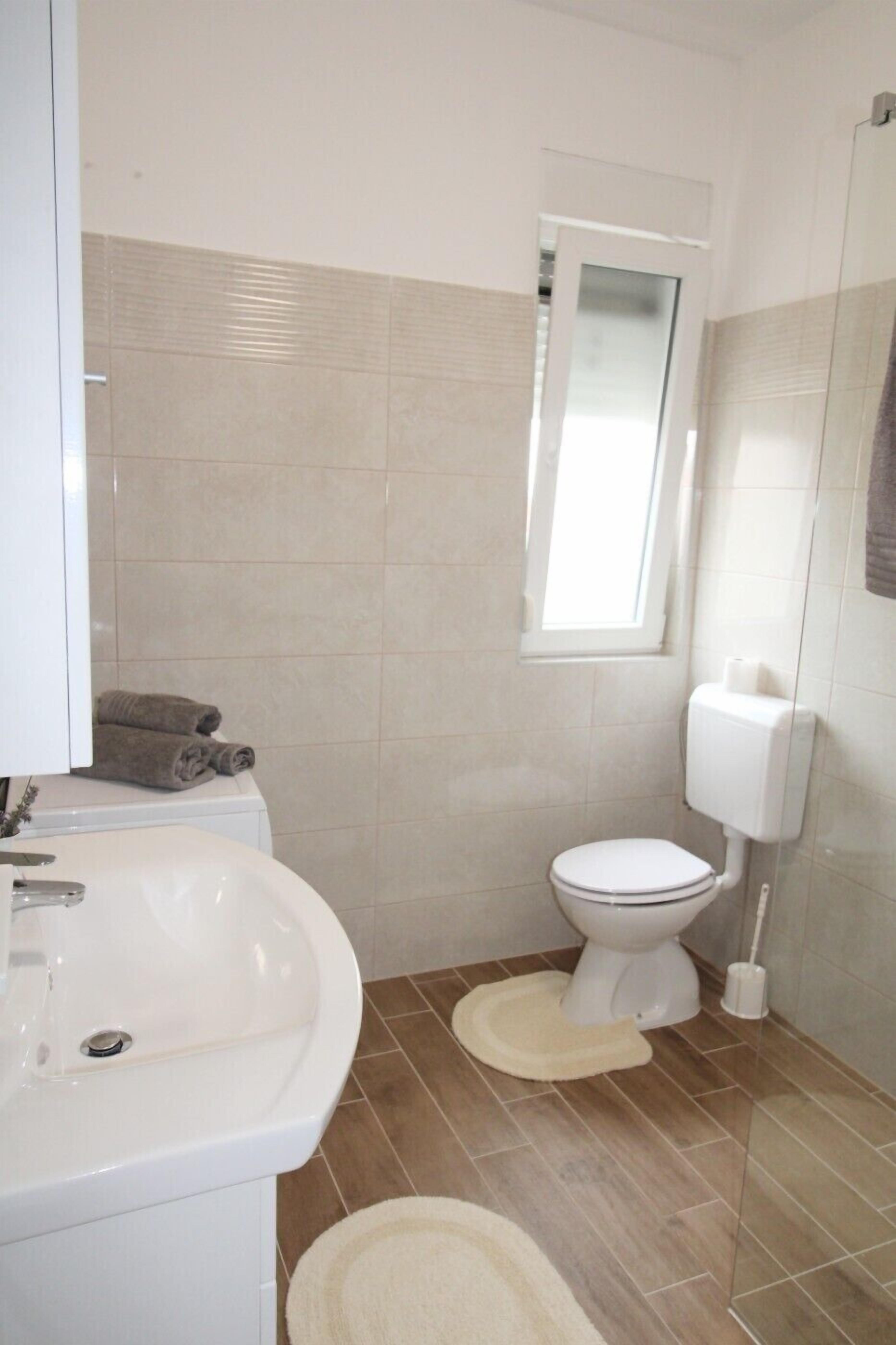 Une salle de bain avec un lavabo blanc, des toilettes et une fenêtre située au-dessus des toilettes.