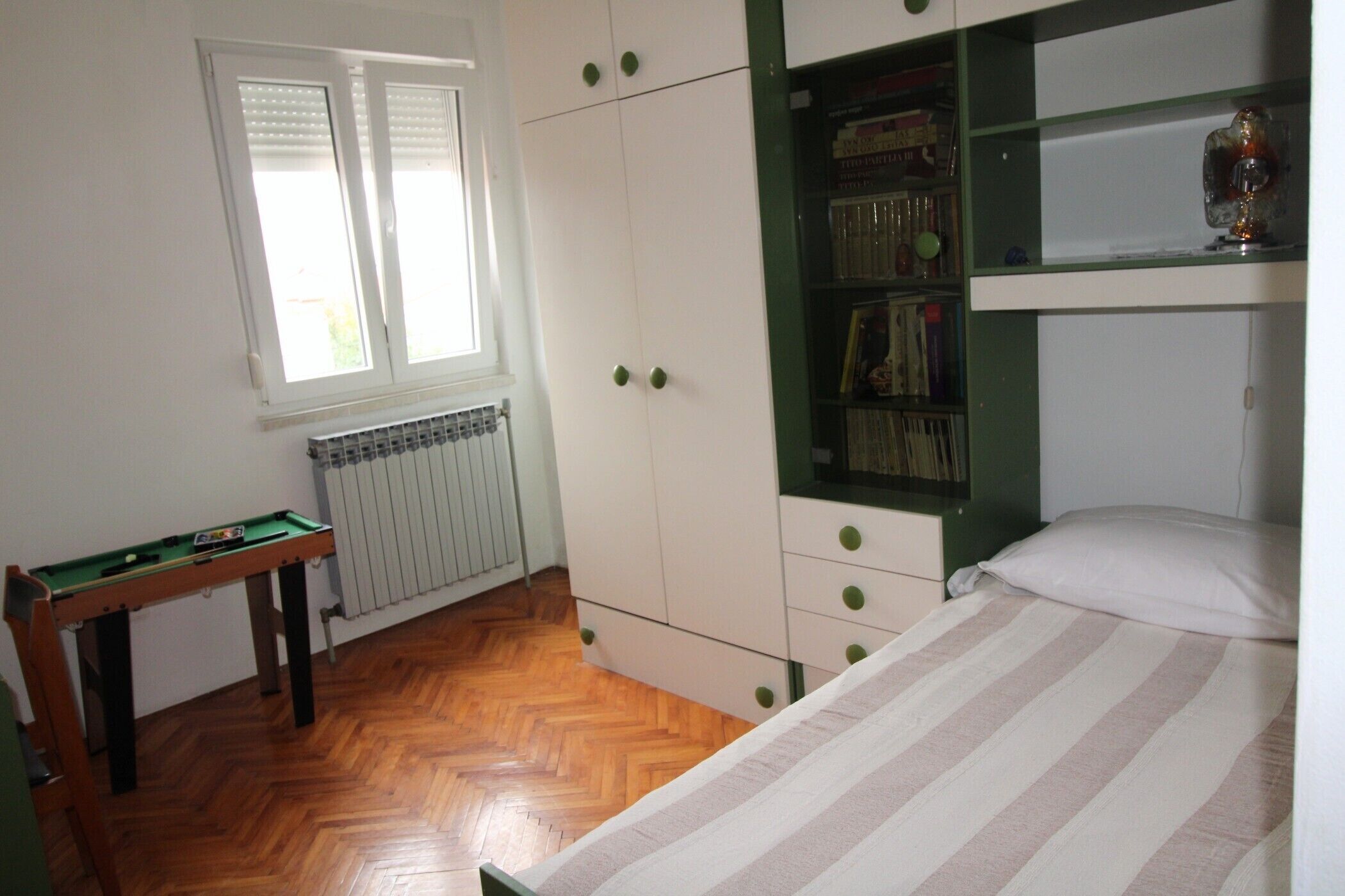 Une pièce comprenant un lit, une bibliothèque verte, un radiateur et une fenêtre avec des stores.