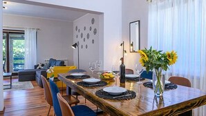 Un comedor con una mesa de madera dispuesta para cuatro personas, rodeada de sillas. Hay un jarrón con girasoles sobre la mesa, y un frutero a la izquierda. La habitación tiene un diseño moderno con un sofá y una lámpara al fondo.