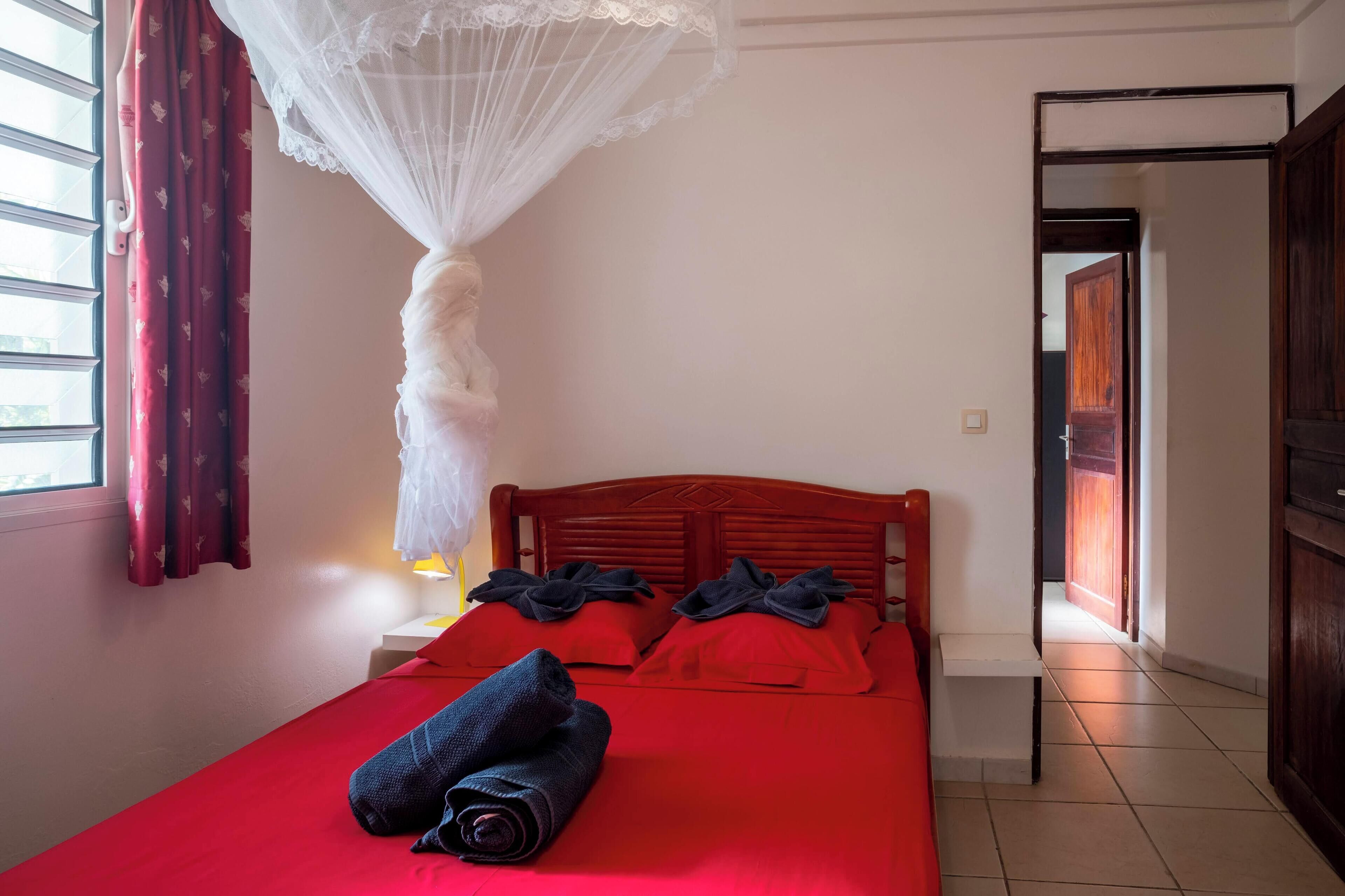 Une chambre d’hôtel avec un lit, une literie rouge, un baldaquin blanc et une vue sur le couloir.