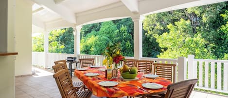 An outdoor covered patio with a dining table set for six, surrounded by wooden chairs, a vase with flowers, and a bowl of fruit.