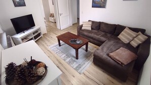 Uma sala de estar com um sofá marrom, uma mesa de centro, uma TV sobre um suporte e uma porta que dá para outro cômodo.