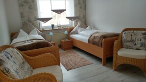 Un dormitorio con dos camas individuales, una mesita de noche de madera, una mesilla con lámpara y una silla de mimbre, además de un ventanal con cortinas.