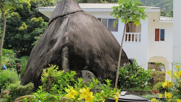 A large rock formation in front of a white building with a balcony, surrounded by lush greenery and yellow flowers.